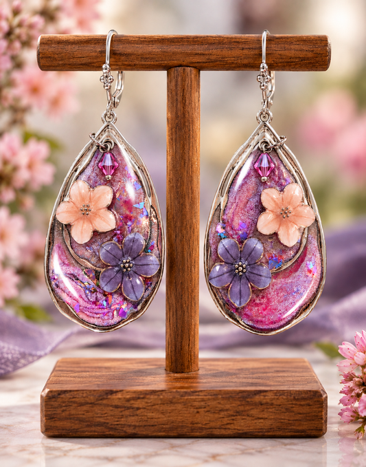 A pair of teardrop-shaped dangling earrings displayed on a wooden stand. The earrings have silver hooks and frames with a pink and purple marbled resin body, featuring a peach flower, a purple flower, and a small purple bead.