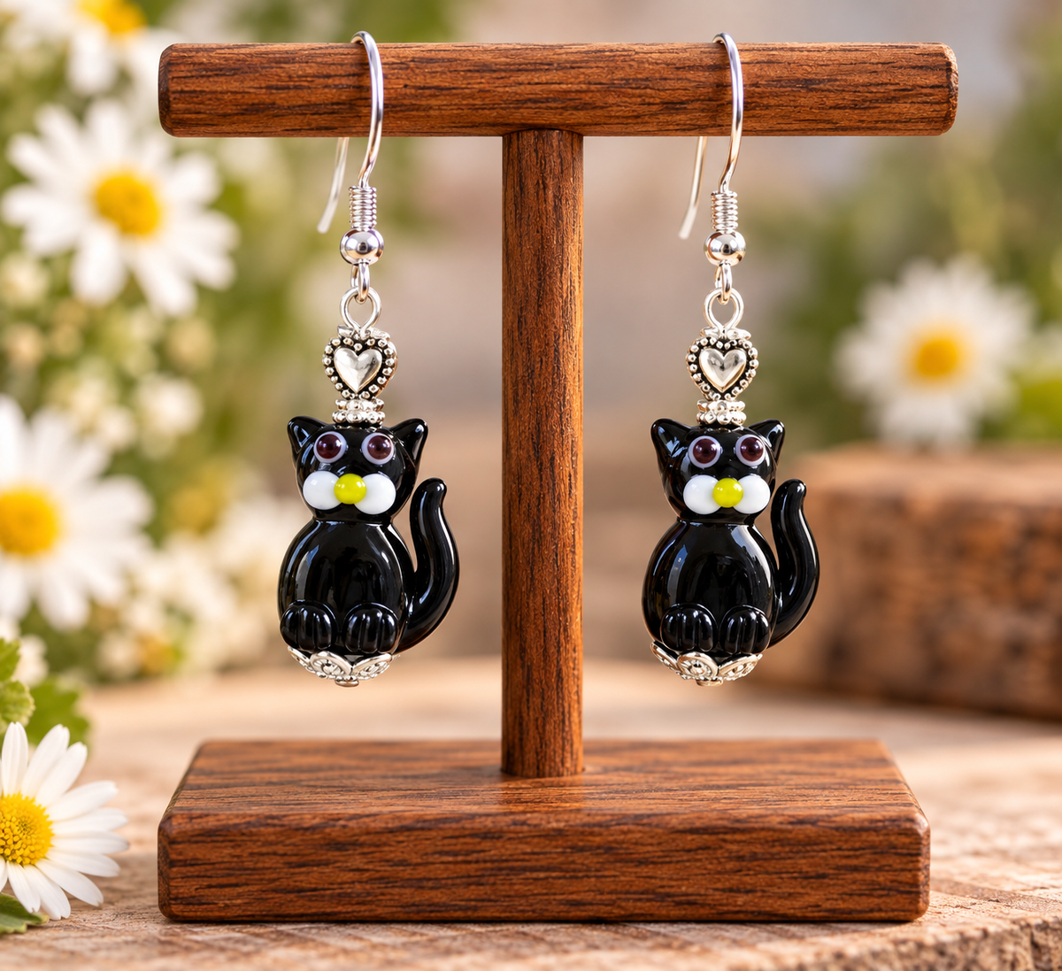 A pair of dangling cat earrings displayed on a wooden stand. Each earring features a glossy black cat charm with white and yellow facial details, a small silver heart, and silver fishhook wires.
