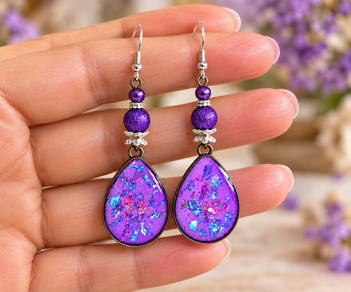 A pair of handmade dangle earrings held in a hand. The earrings feature teardrop-shaped purple resin drops filled with iridescent glitter, accented by purple beads and small clear crystals on silver hooks.
