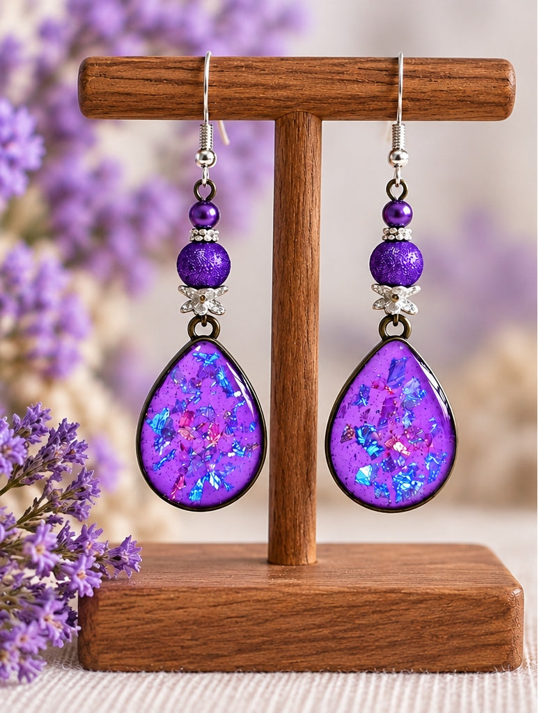A pair of dangling earrings displayed on a wooden T-stand. Each earring has silver hooks, purple beads, and a teardrop-shaped purple iridescent stone.