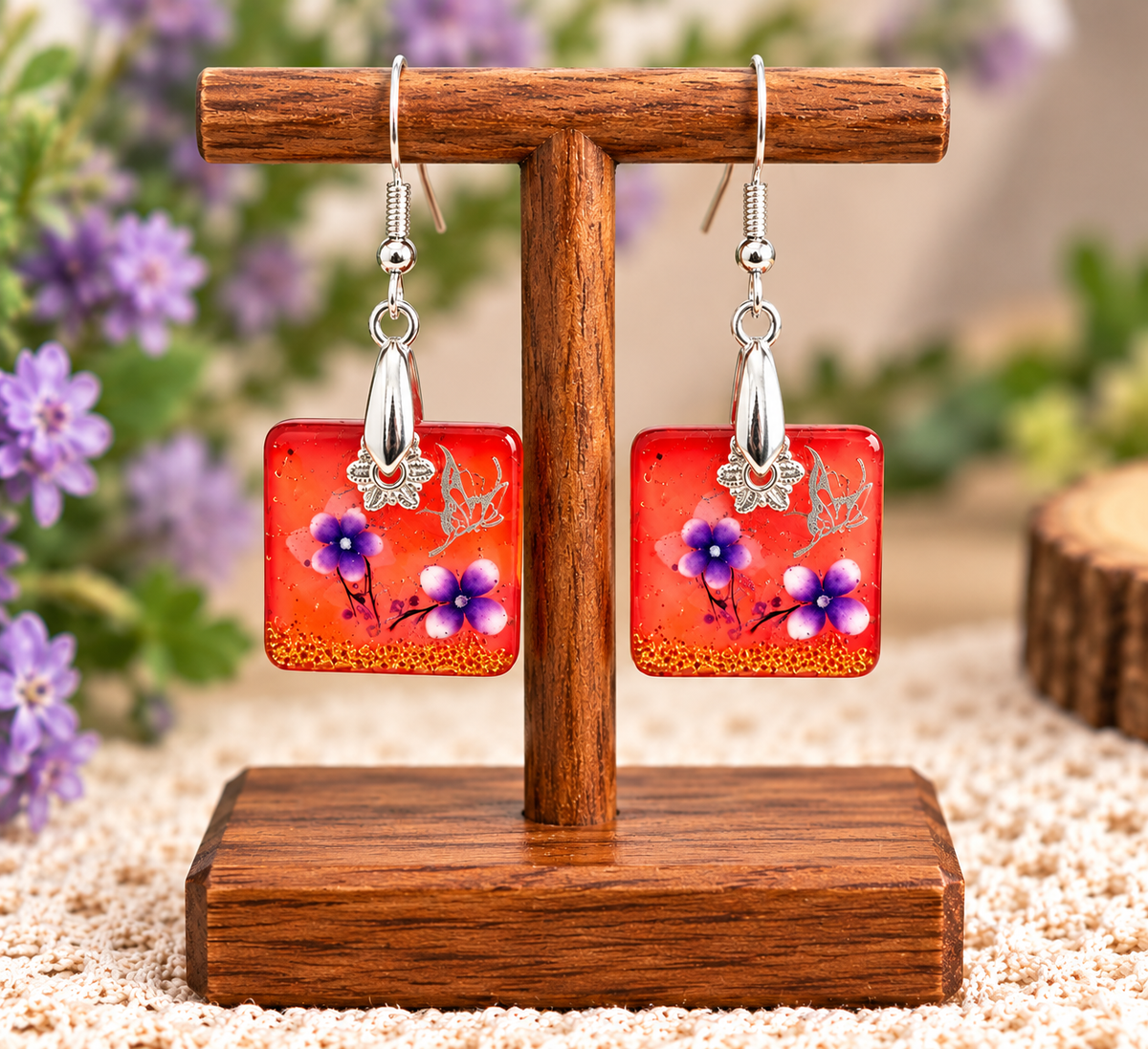 A pair of square dangling earrings displayed on a wooden stand. Each earring has a bright orange-red square pendant with purple and white flowers, delicate green leaves, and small gold glitter details, hanging from silver fishhook wires with silver butterfly charms.