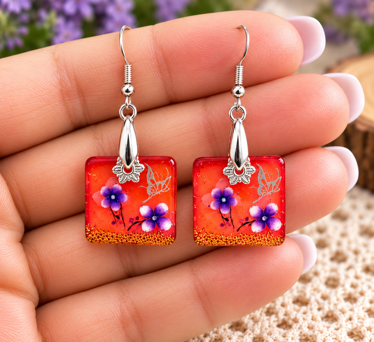 A pair of square glass drop earrings held in a hand. The earrings feature a vibrant orange-red background filled with purple flowers, blue centers, and gold glitter, suspended from silver fishhook wires with silver butterfly charms.