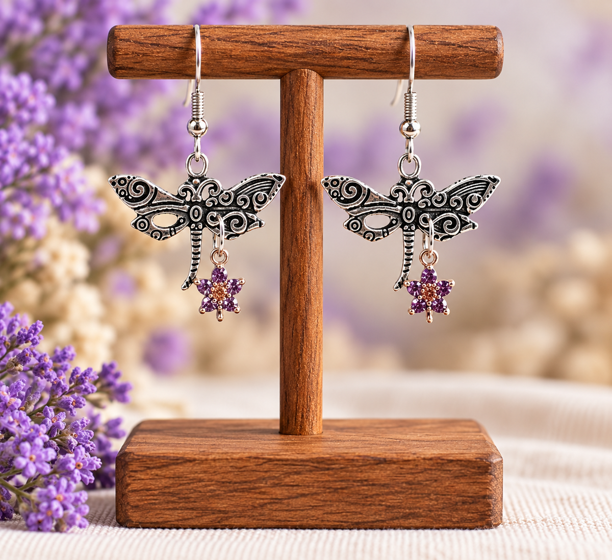 A pair of silver-toned dragonfly dangle earrings with purple flower charms, displayed on a wooden T-stand against a soft, blurred background with purple flowers.