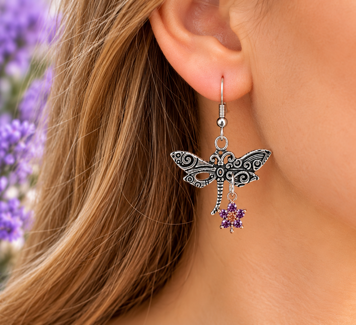 A close-up of a person's ear wearing a dangling dragonfly earring. The earring is made of silver-toned metal with black oxidized detailing in the dragonfly wings and body, and has a small purple flower charm dangling below.