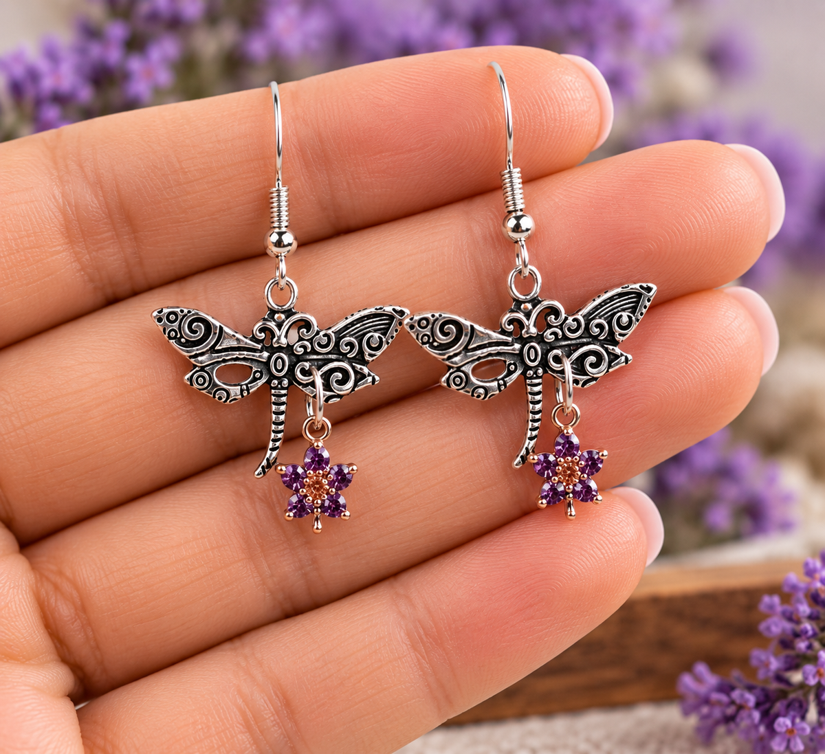 A pair of silver dragonfly dangle earrings with purple crystal flower charms, held on a person's fingers against a blurred background of purple flowers.