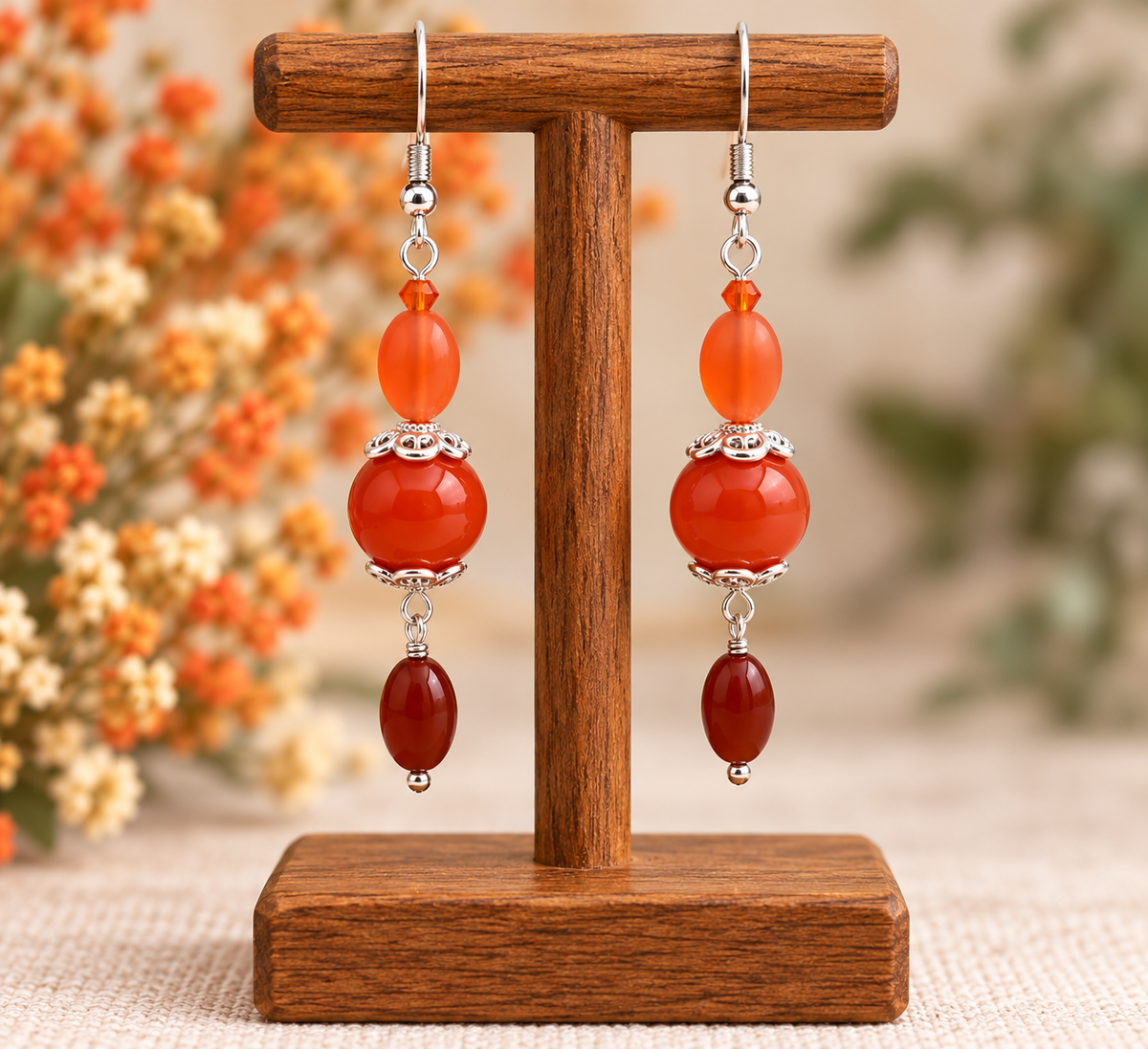 A pair of dangling earrings displayed on a wooden stand. Each earring has silver-tone hooks, a small silver bead, a translucent orange bead, a larger polished orange-red bead, a silver spacer, and a dark reddish-brown bead at the bottom.