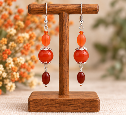 A pair of dangling earrings displayed on a wooden stand. Each earring has silver-tone hooks, a small silver bead, a translucent orange bead, a larger polished orange-red bead, a silver spacer, and a dark reddish-brown bead at the bottom.