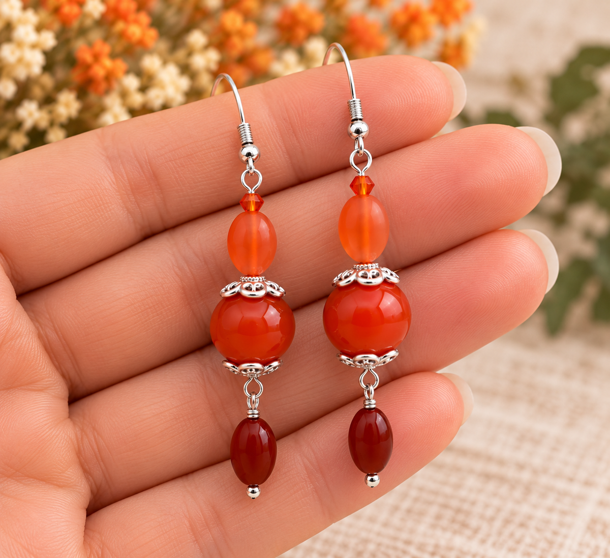 A pair of dangling earrings with carved orange and red stone beads and silver-toned metal hardware, held in a person's hand against a softly blurred background with dried flowers.