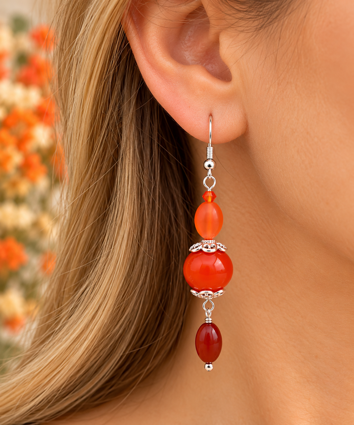 A close-up of a woman's ear wearing a long dangling earring made of orange and red beads with silver-toned metal accents, against a softly blurred background.