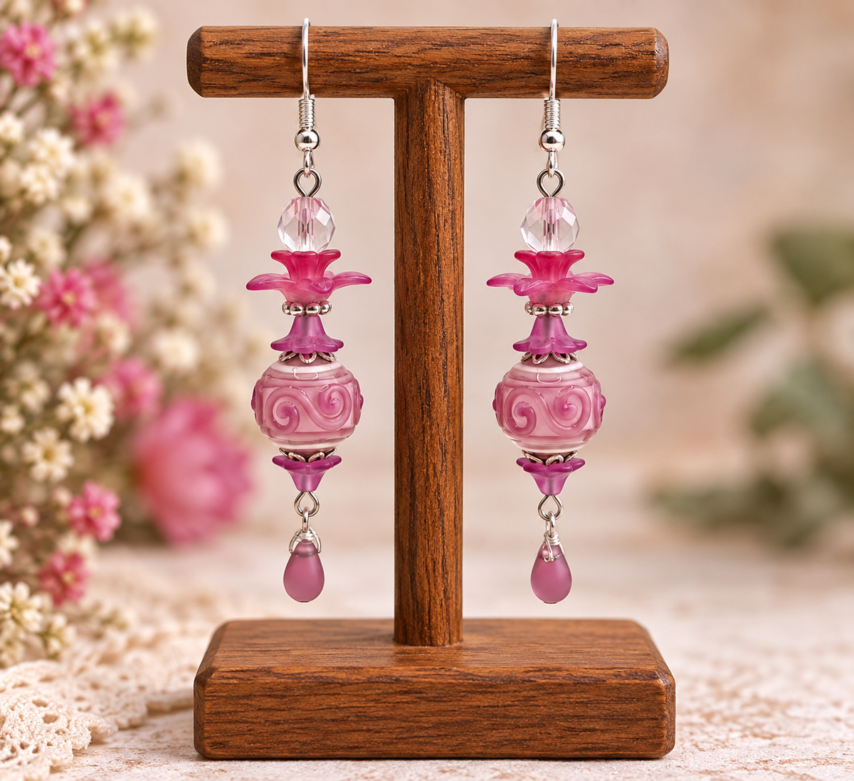 A pair of dangling earrings displayed on a wooden stand. Each earring has silver-tone hooks, a clear faceted bead, a bright pink flower-shaped bead, a pink swirled oval bead, smaller pink floral beads, and a dangling pink teardrop bead.