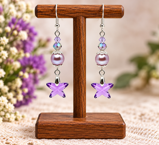 A pair of dangling earrings displayed on a wooden stand. Each earring has silver hooks, iridescent round beads, a lavender pearl-like bead, a faceted crystal, and a purple star-shaped crystal.