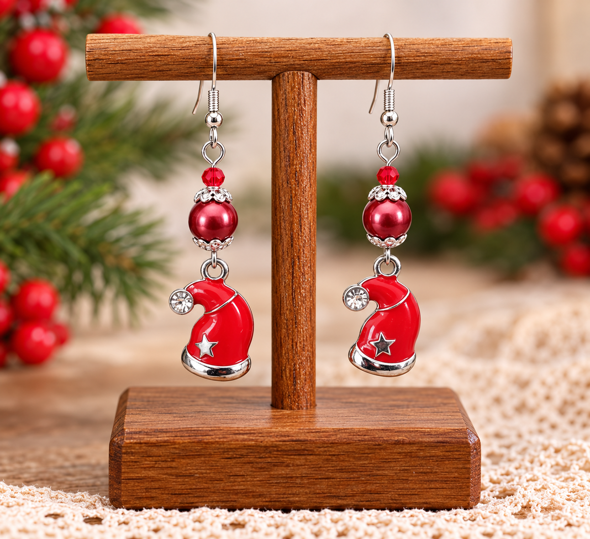 A pair of festive dangle earrings displayed on a wooden stand. Each earring features a silver fishhook, a small red bead, a round red pearl-like bead with silver crystal trim, and a dangling red Santa hat charm with a silver star and silver border.