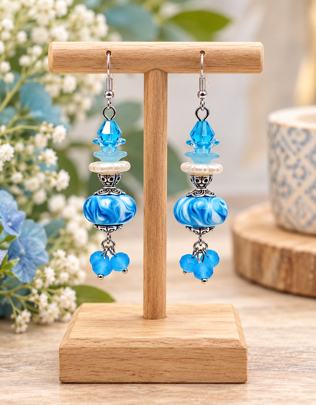 Blue handmade beaded dangle earrings displayed on a natural wooden jewelry stand, featuring aqua crystal beads, pearl accents, marbled blue beads, silver-tone details, and dangling blue bead clusters.