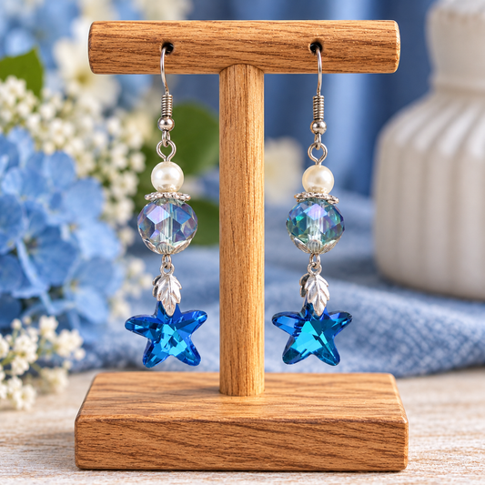 A pair of dangling earrings displayed on a wooden stand. Each earring has a silver fishhook, a white pearl bead, a faceted iridescent blue bead, a small silver leaf charm, and a bright blue star-shaped drop.