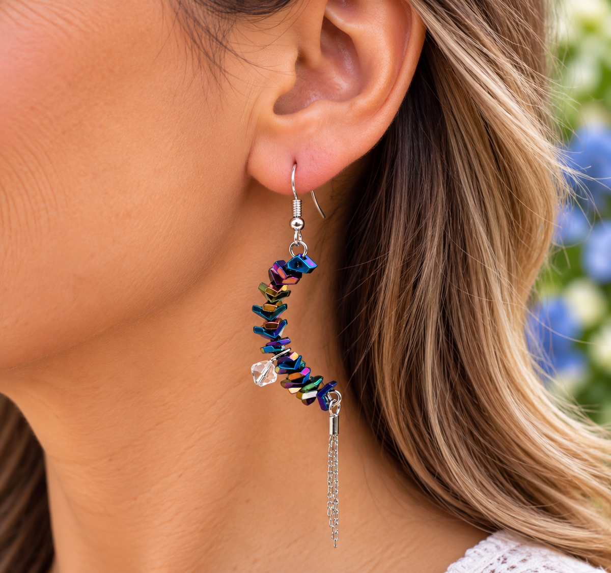 A close-up of a woman's ear wearing a dangling earring made of small iridescent blue, purple, and green beads with a clear heart-shaped crystal and a silver chain tassel.