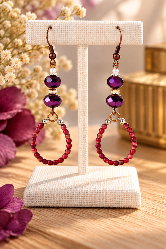 A pair of drop earrings displayed on a cream fabric jewelry stand. The earrings feature faceted purple beads, small gold spacer beads, and dangling beaded loops in deep red.