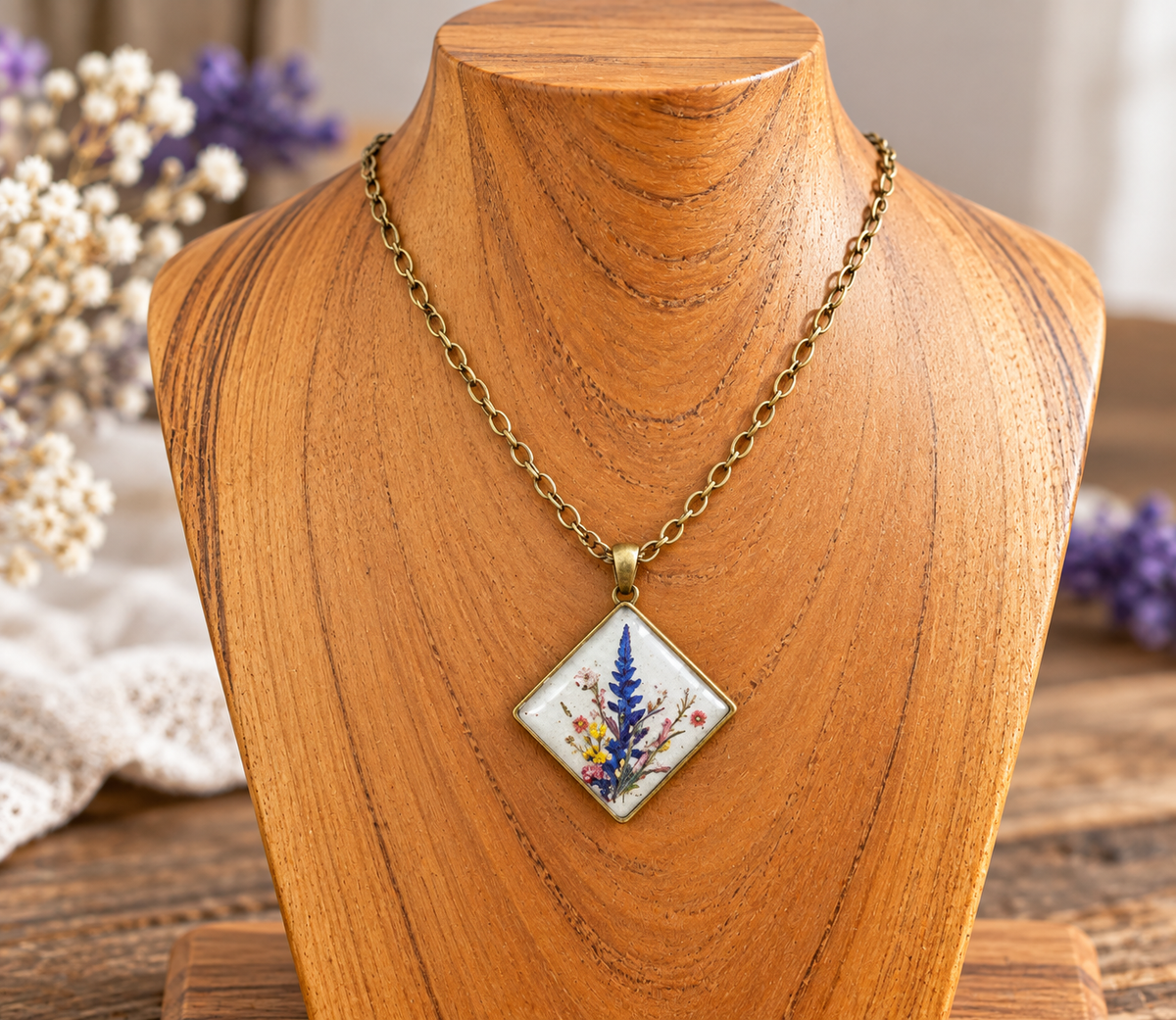 A gold-tone chain necklace with a square pendant featuring a white background and pressed botanical elements in blue, yellow, and pink, displayed on a wooden bust.