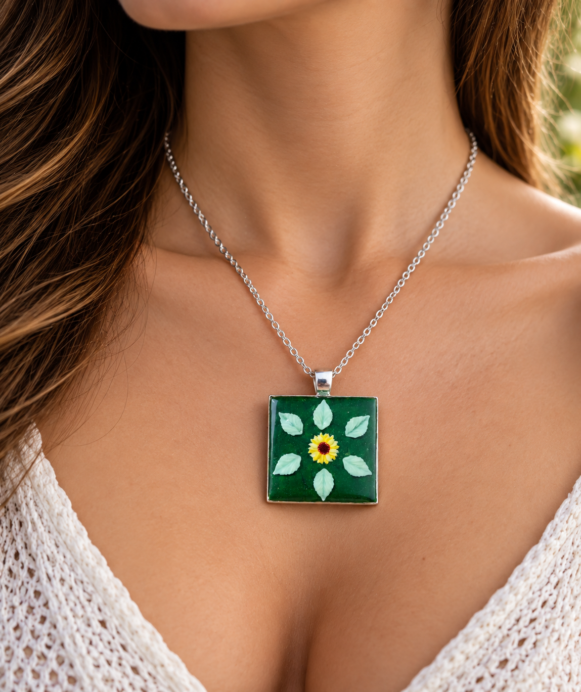 A silver chain necklace with a square pendant featuring a green background and a hand-painted style sunflower with white leaves, worn by a model.