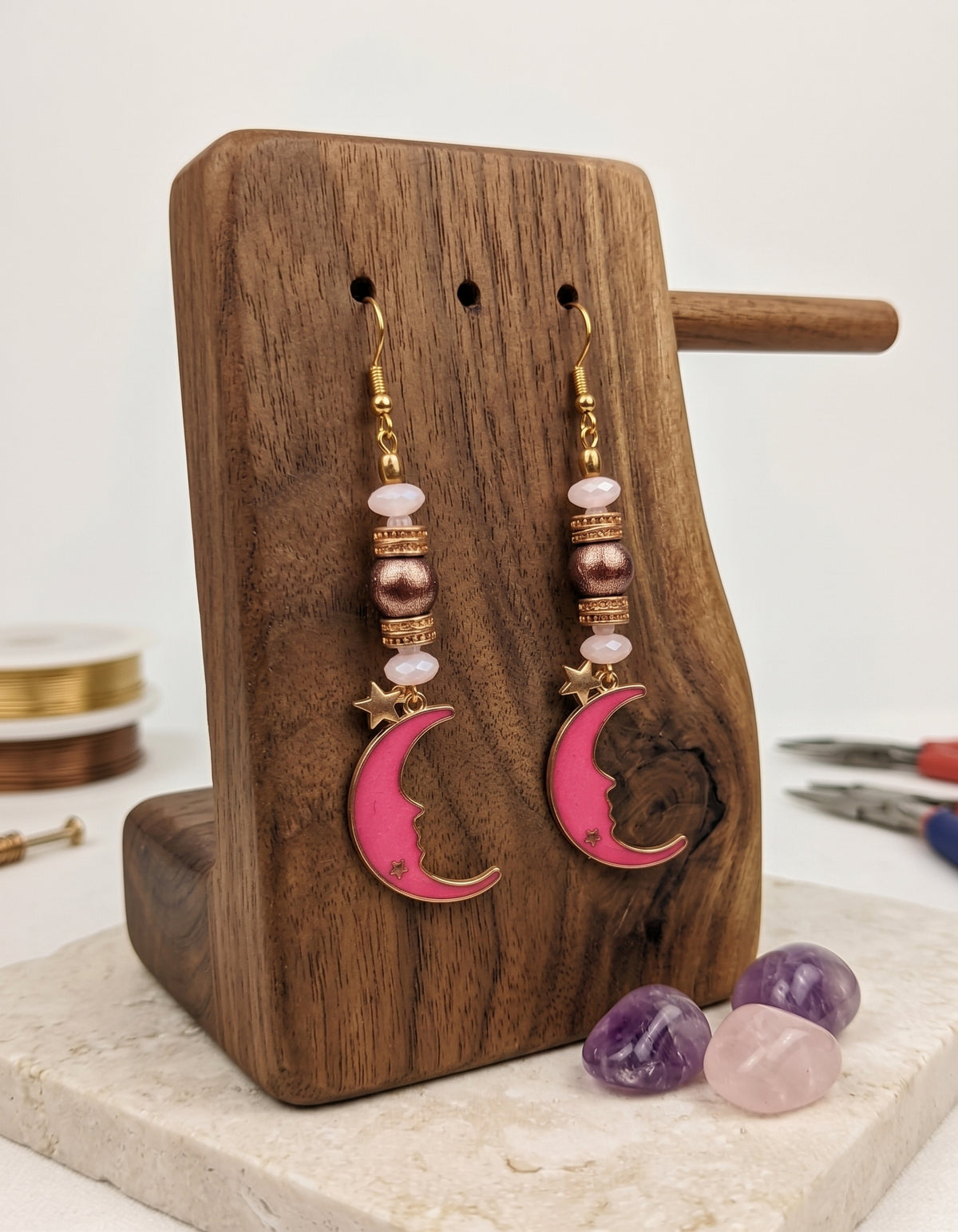 A pair of handmade dangle earrings with pink crescent moon charms, gold-toned hooks, and pink and metallic beads, displayed on a wooden stand.