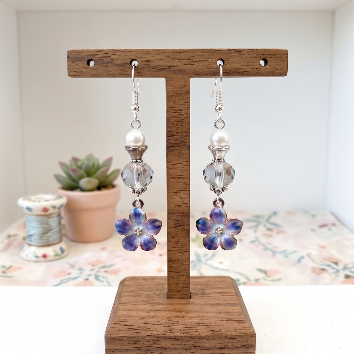 A pair of dangling earrings displayed on a wooden T-stand. Each earring has a silver fishhook, a white pearl bead, a faceted clear crystal bead, and a dangling flower charm with iridescent blue-purple petals and a small clear center.