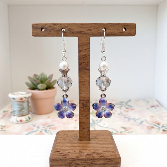 A pair of dangling earrings displayed on a wooden T-stand. Each earring has a silver fishhook, a white pearl bead, a faceted clear crystal bead, and a dangling flower charm with iridescent blue-purple petals and a small clear center.