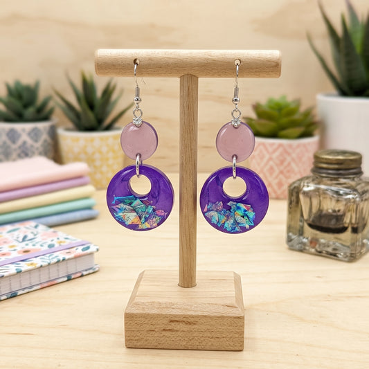 Purple and pink dangle earrings with colorful mosaic inlay, displayed on a wooden stand on a light wood table with small plants and fabric in the background.
