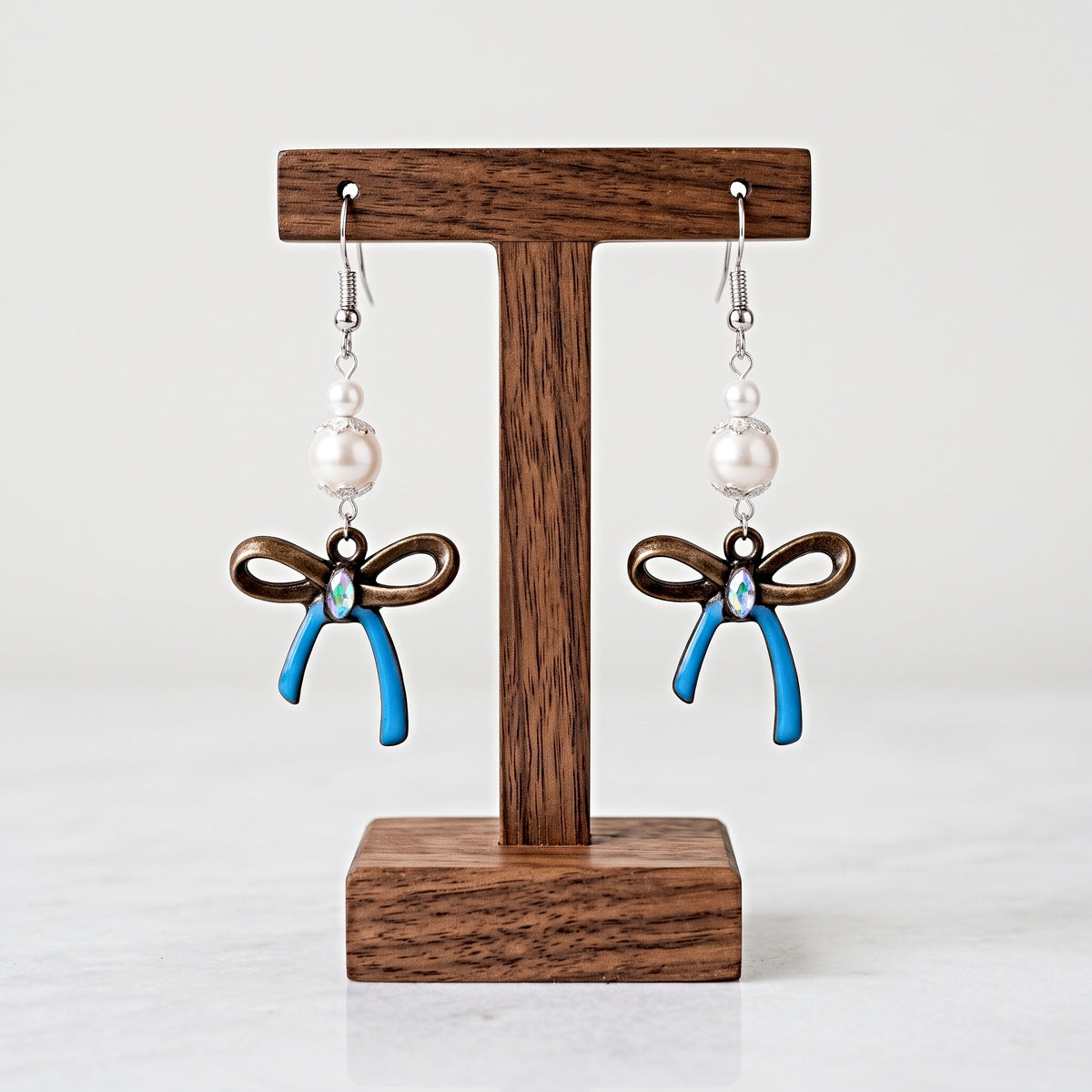 A pair of dangling earrings displayed on a wooden T-stand against a light gray background. Each earring has a silver fishhook, a white pearl bead, a dark brown bow charm, and a blue ribbon-shaped charm with a small clear rhinestone at the center.