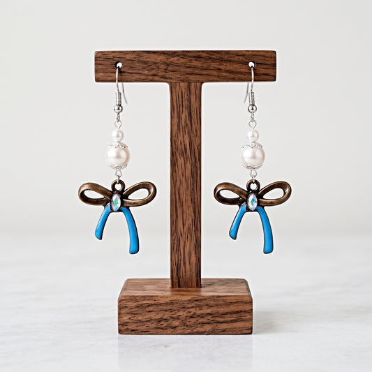 A pair of dangling earrings displayed on a wooden T-stand against a light gray background. Each earring has a silver fishhook, a white pearl bead, a dark brown bow charm, and a blue ribbon-shaped charm with a small clear rhinestone at the center.