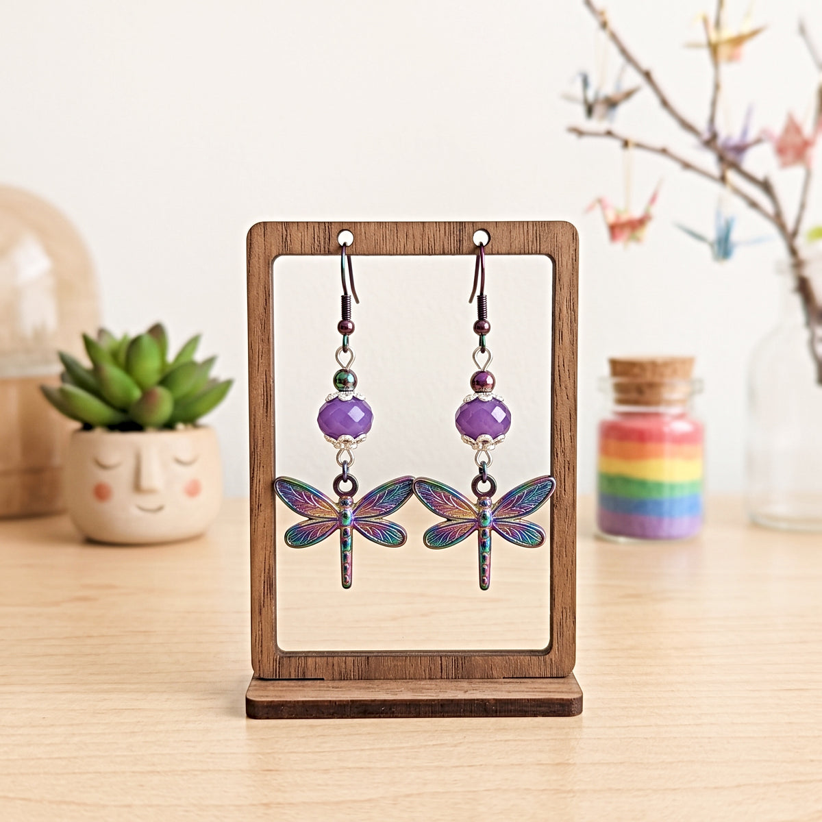 A pair of dangling dragonfly earrings displayed on a small wooden stand on a tabletop. The earrings have purple faceted beads, silver-toned hooks and connectors, and iridescent blue-green dragonfly charms.