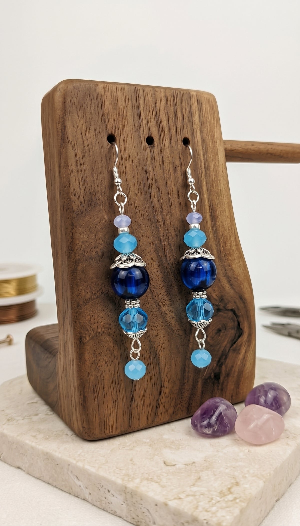 A pair of long beaded dangle earrings displayed on a wooden stand. The earrings feature silver-toned fishhook wires with a series of beads including light blue, dark blue, lavender, and faceted light blue beads, accented by small silver filigree details.