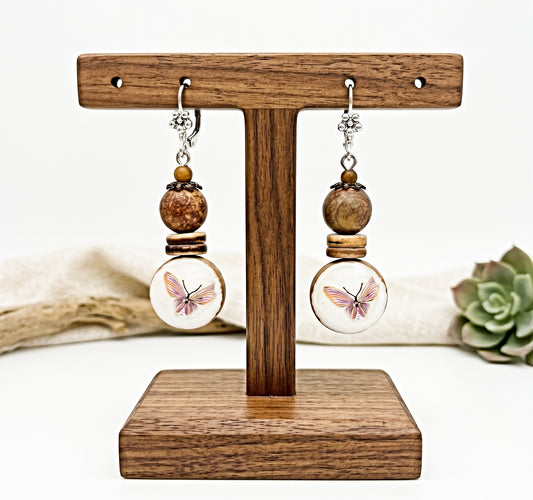 A pair of handmade dangle earrings displayed on a wooden T-stand. The earrings feature silver-toned metal hooks, brown wooden beads, and round white discs with a pink and black butterfly illustration.