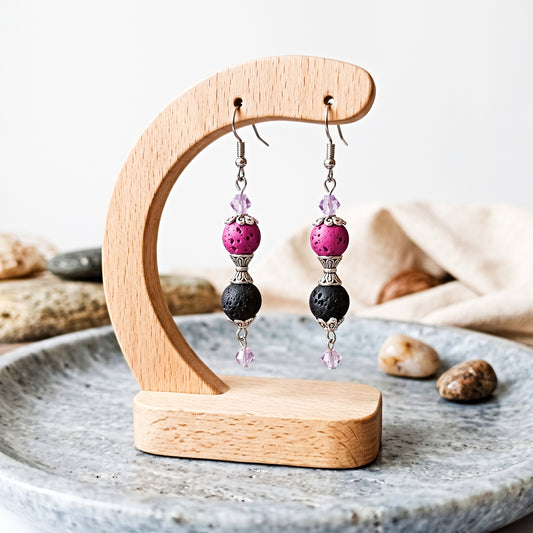 Handmade dangle earrings with pink and black lava beads and light purple crystals, displayed on a wooden stand.