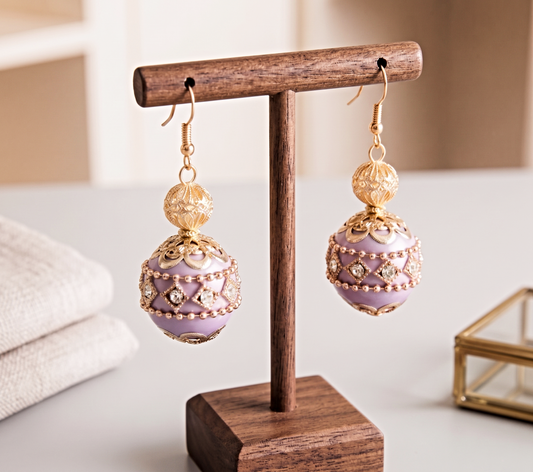 A pair of dangling earrings displayed on a wooden stand. Each earring has a gold-toned fishhook and a round gold-toned bead cap above a lavender-purple spherical drop embellished with gold filigree, rhinestones, and small gold beads.