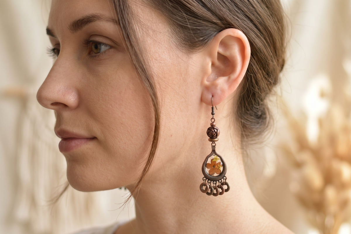 A close-up side profile of a woman wearing a single ornate dangling earring. The earring has a dark bronze-toned metal frame with a warm amber-orange floral inlay and small dangling bead accents.