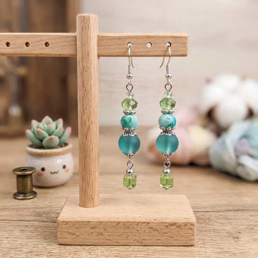 A pair of dangling beaded earrings displayed on a small wooden stand. The earrings have silver hooks with green faceted beads, turquoise-blue round beads, and small silver spacer details, ending in green faceted beads.