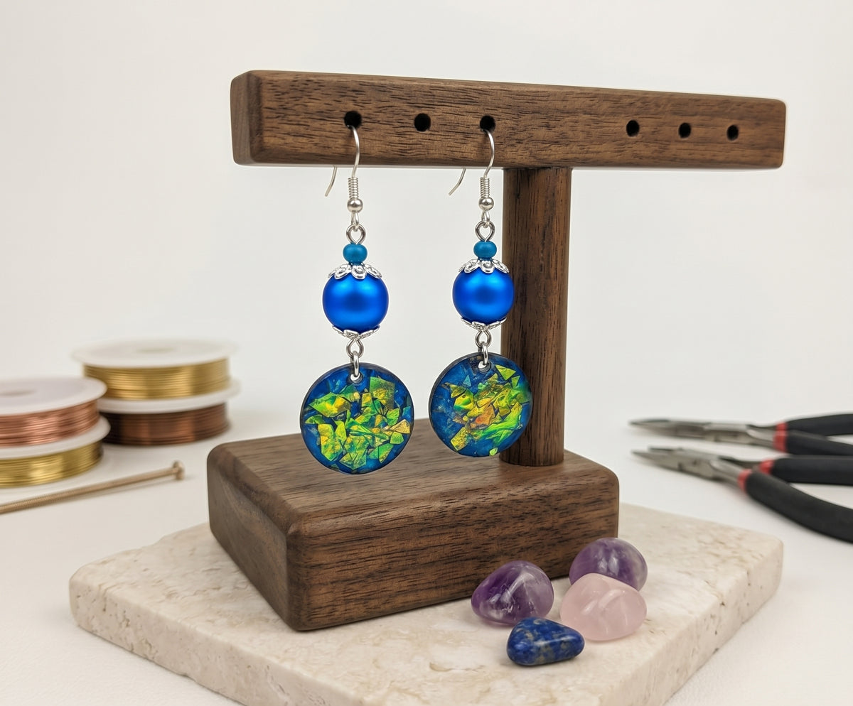Handcrafted dangle earrings displayed on a dark wood T-bar jewelry stand, featuring vibrant royal blue beads, silver filigree bead caps, and round circular charms with green and gold iridescent mosaic flakes