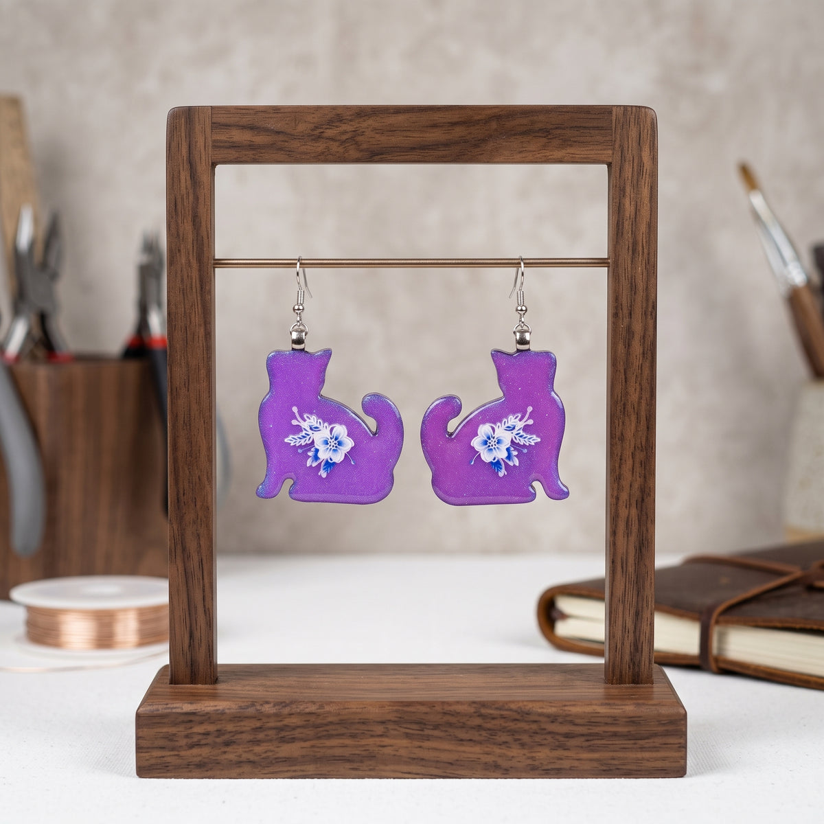 A pair of purple cat-shaped dangle earrings with white floral designs, displayed on a wooden stand.