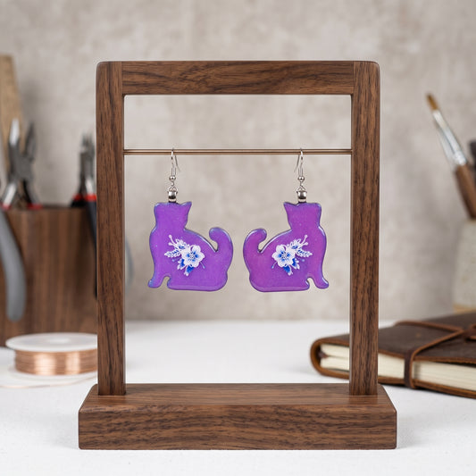 A pair of purple cat-shaped dangle earrings with white floral designs, displayed on a wooden stand.