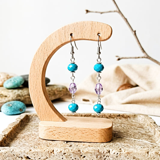 A pair of dangling earrings displayed on a wooden stand. Each earring has silver hooks, turquoise blue beads, and faceted purple crystal links, set against a softly blurred rustic background with stones and branches.