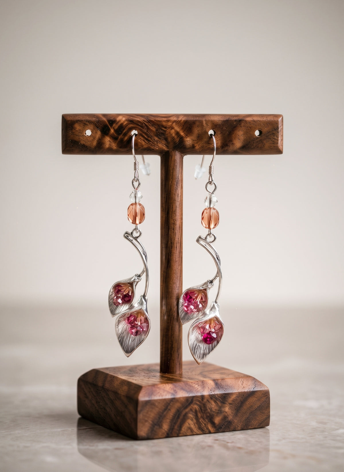 A pair of dangling earrings displayed on a wooden stand against a soft neutral background. The earrings feature silver-toned metal hooks and curved stems with small peach-orange beads and teardrop-shaped pink gemstones with silver leaf-like accents.