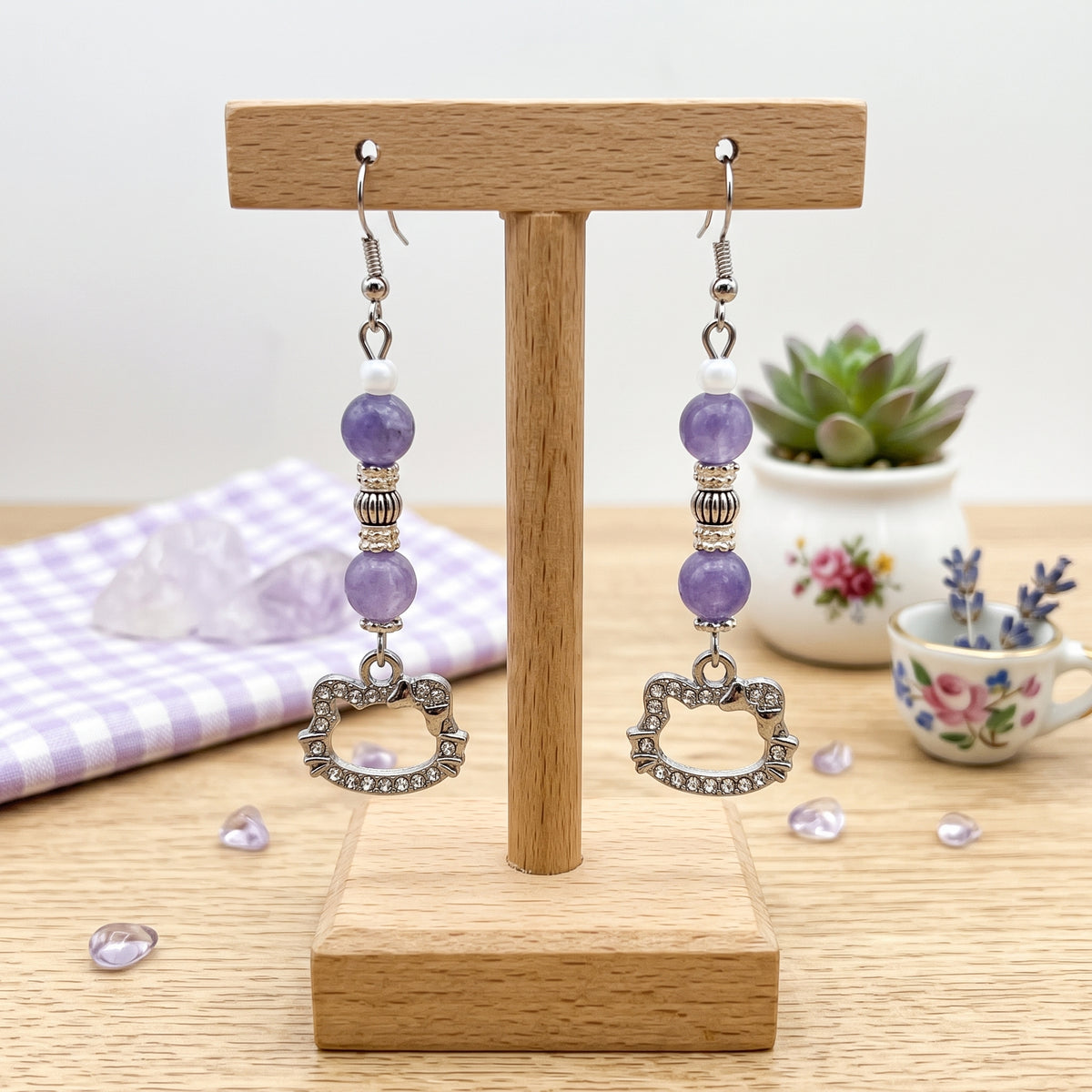 A pair of dangling earrings displayed on a wooden T-stand. Each earring has silver-tone hooks, purple round beads, a small silver spacer bead, a clear faceted bead, and a silver heart-shaped charm set with small clear stones.