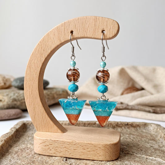 A pair of dangling earrings displayed on a curved wooden stand. Each earring has silver hooks, turquoise blue beads, a brown marbled bead, a round turquoise bead, and a triangular drop with a blue top half and an orange-brown bottom half.