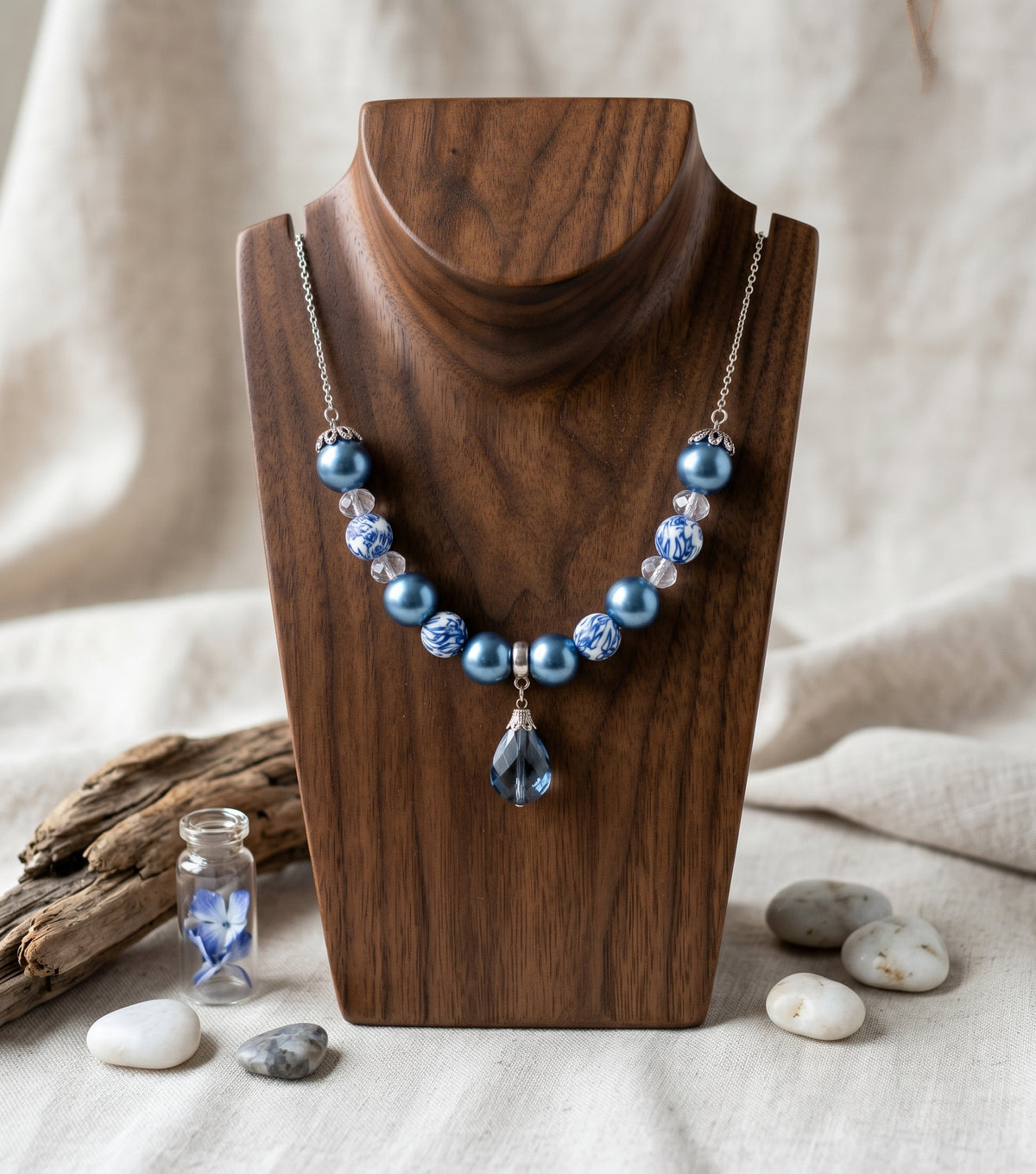 A beaded necklace with light blue pearls, textured blue beads, and a teardrop pendant, displayed on a dark wood stand against a neutral linen background with decorative stones and driftwood.