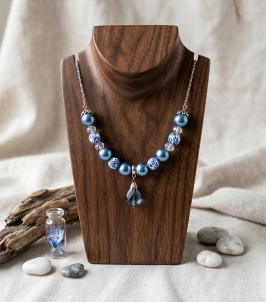 A beaded necklace with light blue pearls, textured blue beads, and a teardrop pendant, displayed on a dark wood stand against a neutral linen background with decorative stones and driftwood.