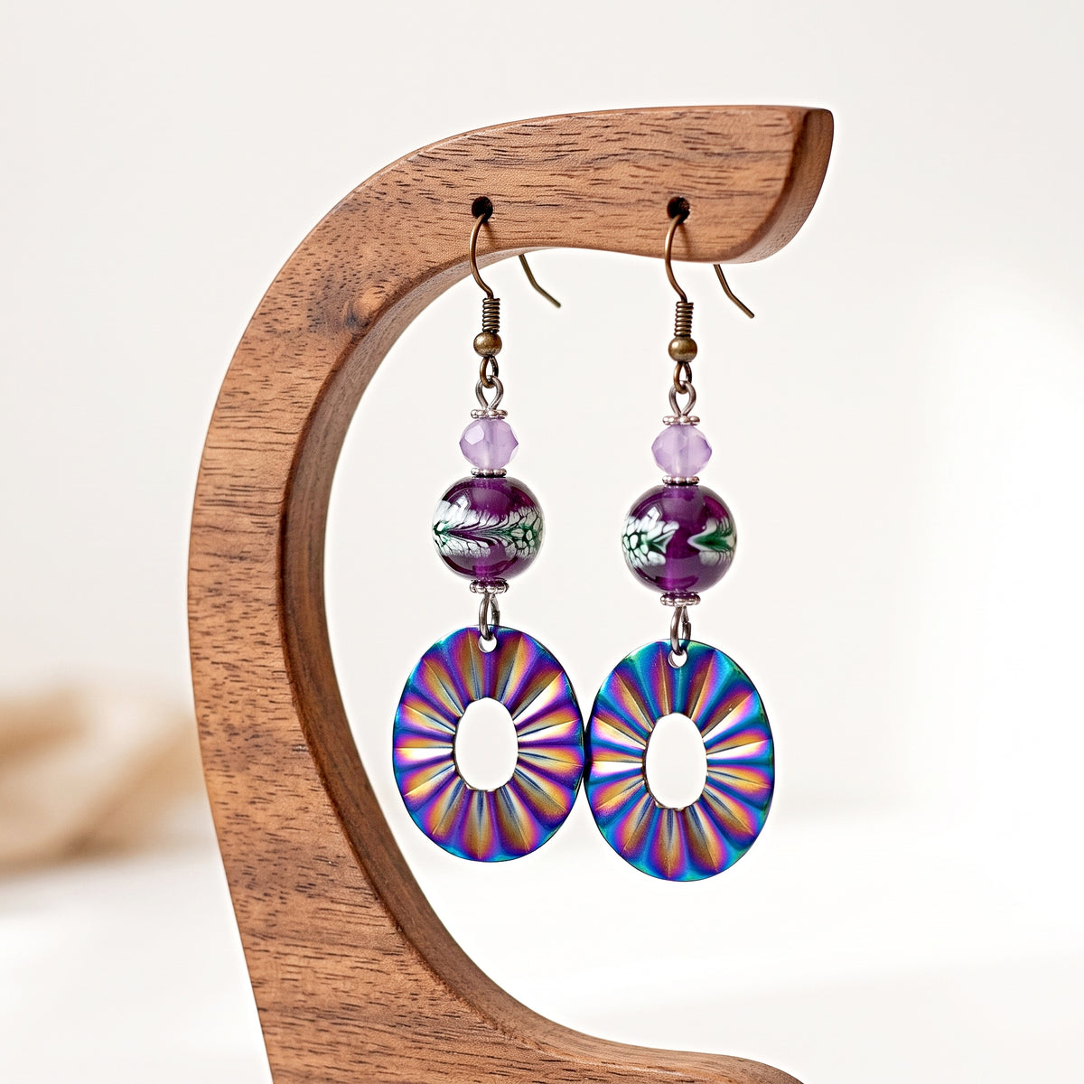 A pair of dangling earrings displayed on a wooden stand against a soft light background. Each earring has a purple marbled bead, a small lavender spacer bead, and a large circular disc charm with a multicolor sunburst pattern in purple, blue, pink, and yellow.
