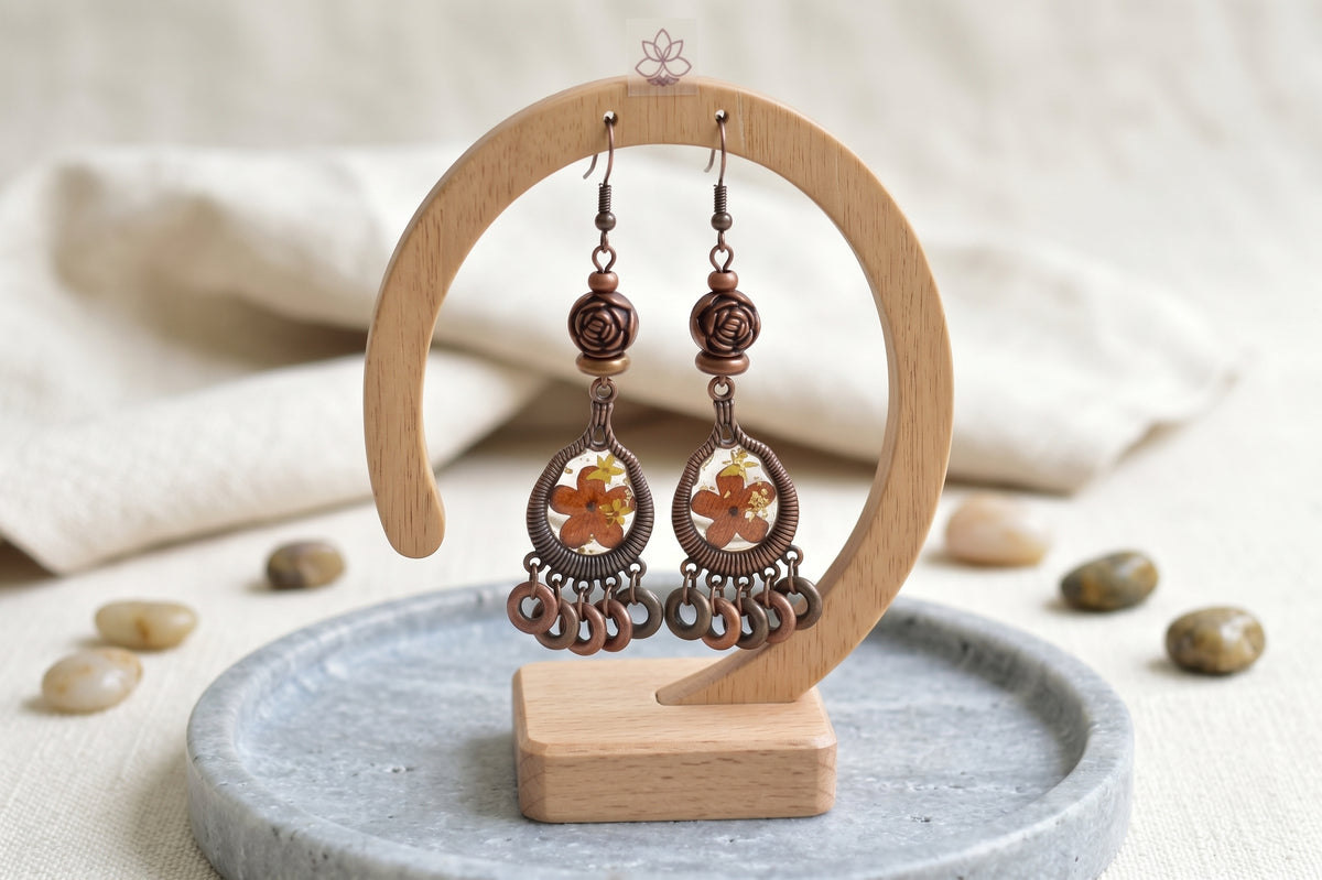 A pair of handmade copper and amber dangle earrings displayed on a wooden stand, set against a soft beige fabric background with small decorative stones.