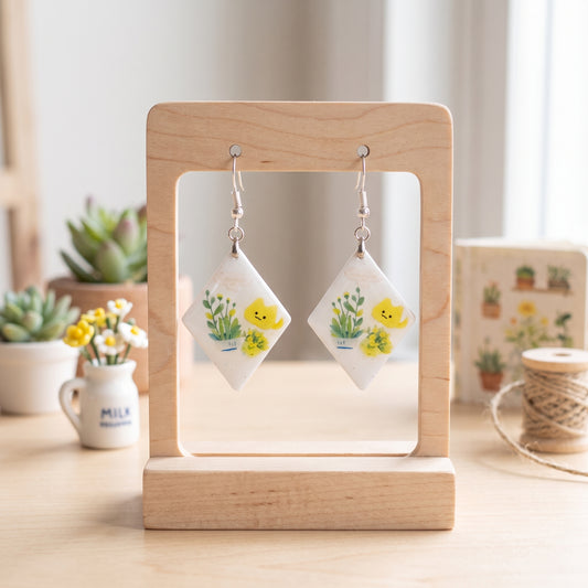 A pair of handmade dangle earrings displayed on a wooden stand. The earrings feature white diamond-shaped pendants decorated with yellow ducklings and green plants, hanging from silver hooks.