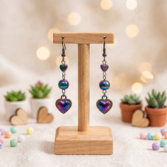 A pair of dangling heart earrings with iridescent purple and blue heart charms, displayed on a small wooden stand against a softly blurred bokeh background with small potted succulents.
