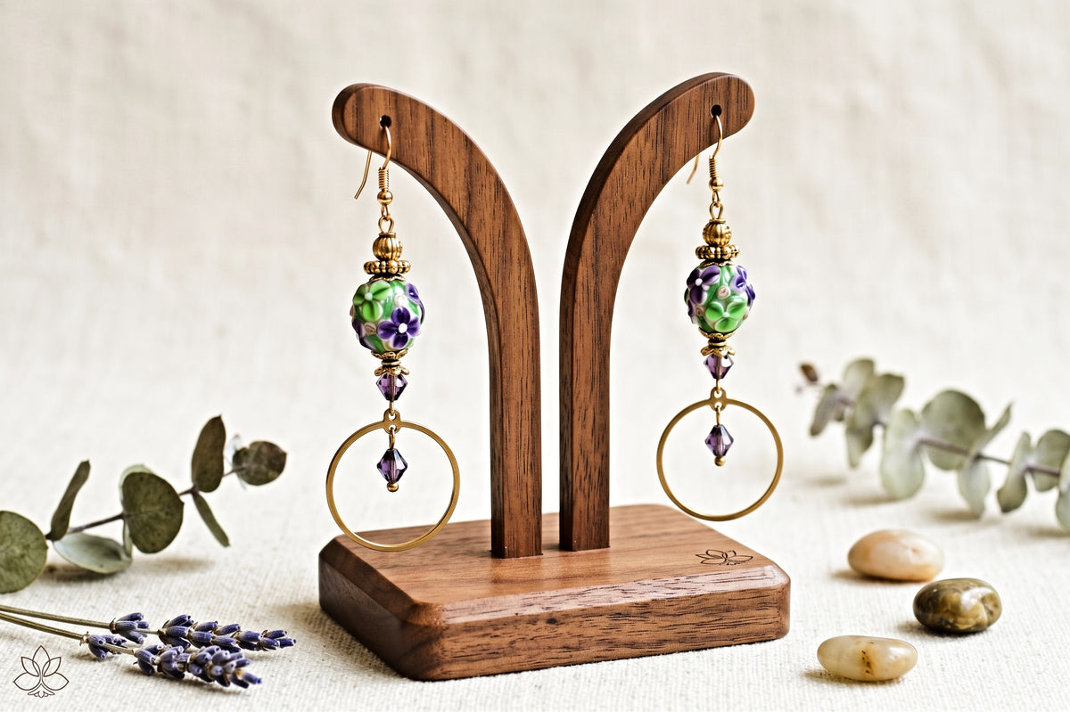 A pair of handmade dangle earrings displayed on a wooden stand. The earrings feature green and purple marbled beads, gold-toned wire hoop details, and small purple crystal accents.