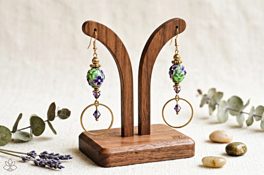 A pair of handmade dangle earrings displayed on a wooden stand. The earrings feature green and purple marbled beads, gold-toned wire hoop details, and small purple crystal accents.